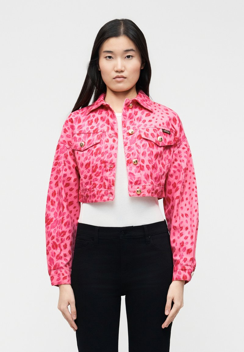 Γυναίκα που φοράει ένα κοντό τζάκετ με ροζ λεοπάρ σχέδιο πάνω από ένα λευκό τοπ και μαύρο παντελόνι, στέκεται μπροστά σε ένα απλό φωτεινό φόντο.