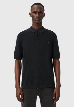 Man draagt een zwart gebreid poloshirt met korte mouwen en een donkere spijkerbroek, staand tegen een effen lichtgrijze achtergrond.