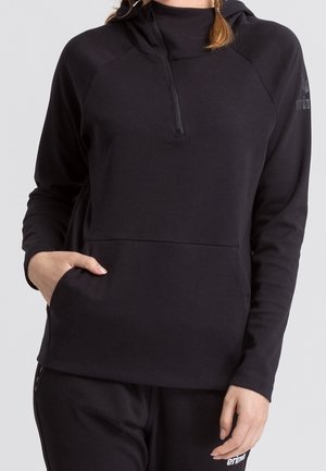 Vrouw draagt een zwarte hoodie met lange mouwen, een halve rits en een voorzak, gecombineerd met bijpassende zwarte broek.