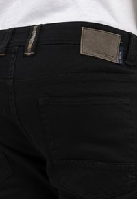 Czarne dżinsy z denimu z prostokątną skórzaną naszywką na pasku z logo marki "camel active". Widoczna tylna kieszeń oraz detale stębnowania.