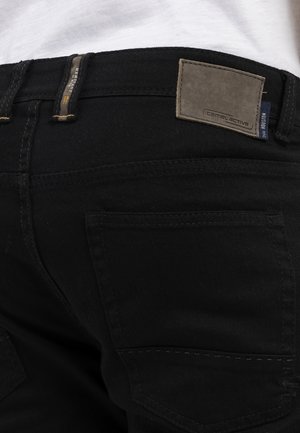 Schwarze Jeans aus Denim mit einem rechteckigen Lederpatch am Bund, das die Marke "camel active" zeigt. Sichtbare Gesäßtasche und Nähte.