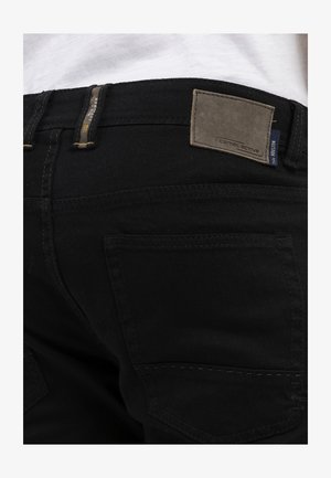 Sorte denimjeans med et rektangulært lædermærke på taljen med mærket "camel active." Synlig baglomme og syninger.