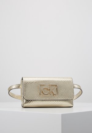 Złota metaliczna torebka na pasek o fakturze, z przednią klapką i złotym logo CK, prezentowana na białej powierzchni na tle prostego szarego tła.