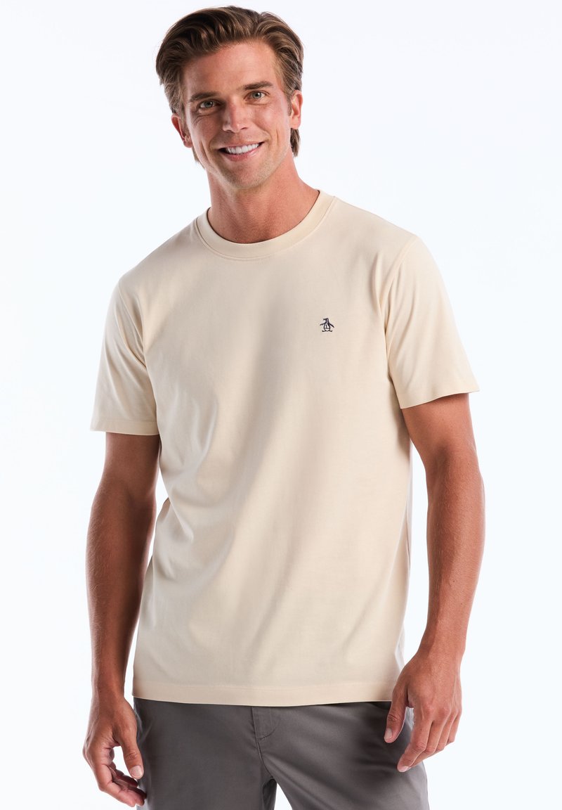 T-shirt beige à manches courtes en tissu lisse, avec un petit logo de pingouin marine sur la poitrine, un col rond et une coupe décontractée.