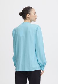 Blusa azzurro chiaro con maniche lunghe, tessuto texturizzato, spalle plissettate e dettaglio sul collo in balze. Abbinata a pantaloni neri.