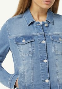 Primo piano della parte superiore del corpo di una donna che indossa una giacca di jeans blu chiaro con bottoni argentati e tasche sul petto, che mostra uno stile informale.