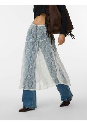 Osoba ubrana w niebieskie dżinsy z przezroczystym, białym koronkowym nakładką w formie spódnicy, czarną bluzkę, brązowe spiczaste buty i niosąca brązową torebkę z frędzlami.