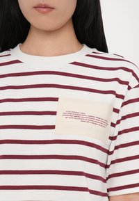 Stribet t-shirt i hvid og maroon med en cremet lomme med trykt tekst. Rund hals og korte ærmer. Blødt bomuldsstof.