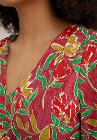 Tkanina w kwiatowy wzór w kolorze czerwonym, żółtym i zielonym z teksturowanym wykończeniem. Posiada dekolt w kształcie V i krótkie rękawy.