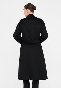 Cappotto nero di lana con maniche lunghe, vita cinturata e lunghezza fino al polpaccio. Presenta un colletto e un design minimalista.