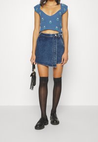 Kvinna som bär en blå blommig kort topp, asymmetrisk denimkjol, svarta knästrumpor, svarta loafers och håller en liten svart handväska.