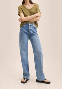 Olijfgroene V-hals T-shirt gecombineerd met lichtblauwe high-waisted wijde jeans en zwarte flip-flops, tegen een neutrale achtergrond.