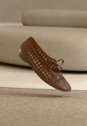 Chaussure plate en cuir tressé marron avec motif découpé et nœud, posée sur un sol neutre moquetté, avec un canapé beige en arrière-plan.