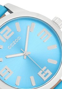 Montre ronde avec un cadran bleu clair, index et aiguilles argentés, et un boîtier argenté. Présente un bracelet bleu clair et le logo de la marque "oozoo".