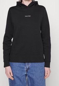 Černá mikina z bavlněné směsi s přední kapsou, na hrudi se bílým logem "Calvin Klein" a ribovanými manžetami a lemem.