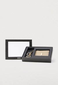 Porta carte in pelle beige con accessori in oro e portachiavi, esposto in una scatola nera con uno scomparto per carta bianco vuoto.