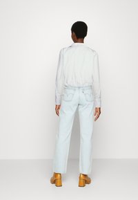Weißes Hemd mit Kragen, hellblaue hochgeschnittene Jeans mit Gesäßtaschen, die mit einer geschwungenen Naht verziert sind, und beigefarbene Absatzschuhe.
