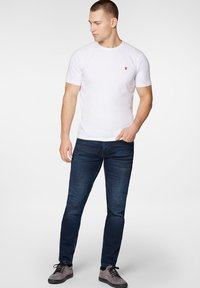 Biała koszulka z krótkim rękawem z małym czerwonym logo, w połączeniu z ciemnoniebieskimi dżinsami slim-fit z przetarciami oraz szarymi trampkami.