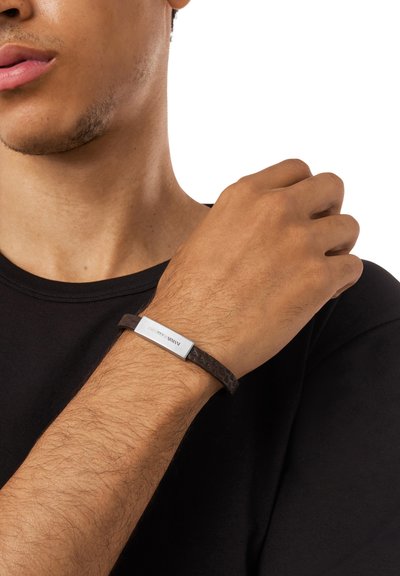 Bracelet en cuir marron avec un design texturé et une plaque rectangulaire en argent gravée "Emporio Armani." Porté au poignet.