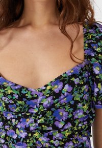 Blommig topp med svart tyg och lila och gröna blommor. Den har en hjärtformad halsringning och puffärmar med sammetsdetaljer.