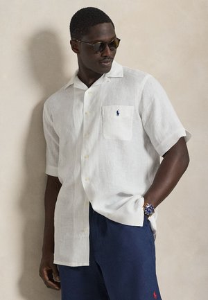 Λευκό πουκάμισο από λινό με κοντά μανίκια, κουμπωτό μπροστινό μέρος και αριστερή τσέπη στο στήθος με μπλε λογότυπο. Συνδυάζεται με σκουρό μπλε σορτς.