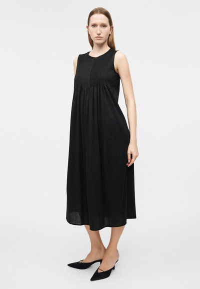 Γυναίκα που στέκεται φορώντας μαύρο μίντι φόρεμα χωρίς μανίκια με πιέτες και μαύρα παπούτσια με χαμηλό τακούνι και μυτερή μύτη, σε απλό φόντο.