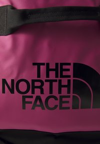 Pink fabric duffel bag with a black logo "THE NORTH FACE" and a half-circle design. Textured surface with a sturdy black handle.