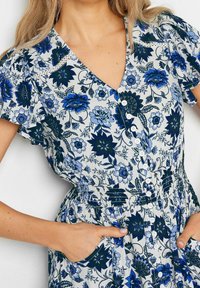 Blommig klänning med blå och marinblå blommor på en ljus bakgrund, V-ringning, korta fladderärmar, knappar och ett elastiskt midjeband med fickor.
