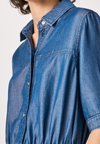 Denim shirt with a button-up collar, short sleeves, and an elastic waist. Made of lightweight blue denim with a subtle sheen and smooth texture.