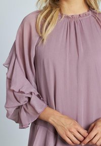 Blusa di un delicato viola chiaro con maniche a sbuffo in tessuto trasparente e scollatura plissettata. Tessuto liscio con una leggera lucentezza. Le mani del modello sono visibili in vita.