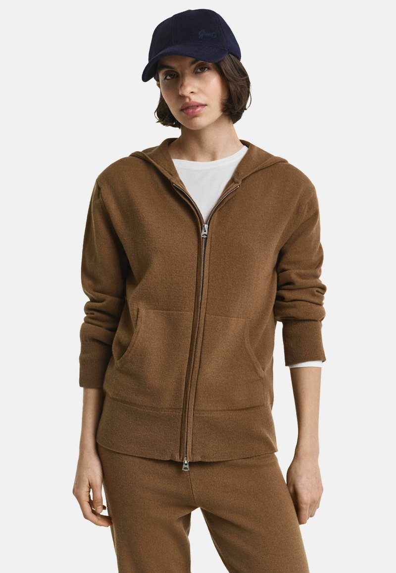 Bruine zip-up hoodie met een capuchon, voorzakken en geribde manchetten, gecombineerd met een marineblauwe pet. Soepele stof, relaxte pasvorm en een minimalistisch ontwerp.