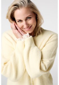 Een zachte, pluizige crèmekleurige cardigan met een ontspannen pasvorm, geribbelde mouwen en kanten rand bij de manchetten, voorzien van een ronde halslijn en knoopsluiting.