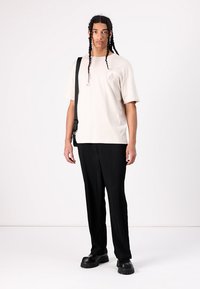 Lys beige bomulds t-shirt med et subtilt logo på brystet, kombineret med sorte plisserede bukser og chunky sorte sko.