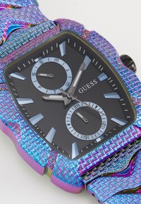 Mehrfarbige Guess-Uhr mit einem strukturierten, schimmernden Armband. Das quadratische schwarze Ziffernblatt verfügt über zwei Nebenziffernblätter und silberne Akzente mit Stundenmarkierungen.