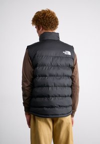 Gilet en duvet noir avec un col haut et un design à coutures horizontales ; présente un petit logo blanc au dos. Porté par-dessus une chemise à manches longues marron.