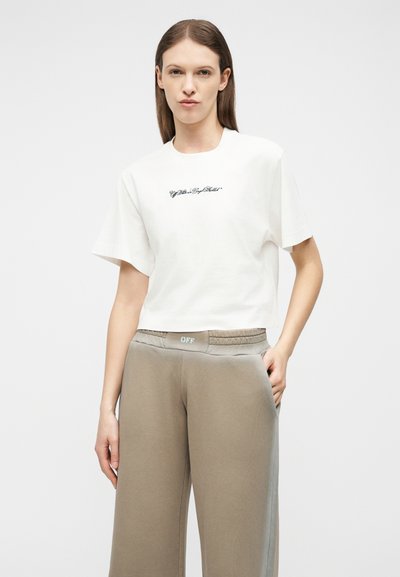 Γυναίκα που φοράει λευκό κοντό μπλουζάκι με κείμενο και μπεζ παντελόνι φαρδιάς γραμμής με ετικέτα «OFF» στη μέση, στέκεται μπροστά σε απλό φόντο.