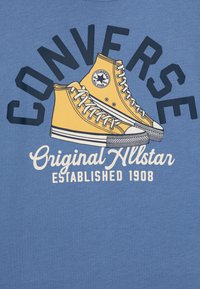 Zila T-kreklu ar dzeltenu augstas klāja kedu grafiku, uz kuras ir melnā krāsā burtus "CONVERSE" un balti "Original Allstar Established 1908".