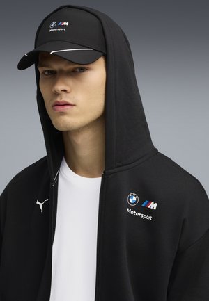 Felpa nera con cappuccio e accenti bianchi, con logo BMW Motorsport, abbinata a un cappellino nero con loghi simili. Texture liscia.