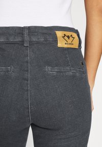 Pantalons gris foncé avec une finition lisse et texturée ; comporte un logo "MOS MOSH" sur une étiquette en cuir beige au-dessus de la taille et deux poches arrière.