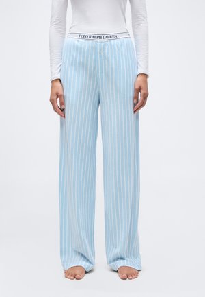 Žena oblečená v svetlomodrých a bielych vertikálne prúžkovaných širokých pyžamových nohaviciach s pásom značky Polo Ralph Lauren a bosými nohami.