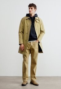 Άντρας που φοράει μπεζ καμπαρντίνα, μαύρο φούτερ με κουκούλα, μπεζ παντελόνι και καφέ παπούτσια στέκεται μπροστά σε άσπρο τοίχο.