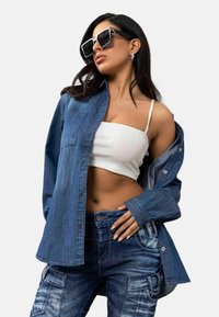 Jeanshemd, hellblau, mit Knöpfen und Brusttasche, über einem weißen gerippten Crop-Top getragen. Blaue Cargo-Jeans mit Taschen und ausgewaschenen Akzenten.