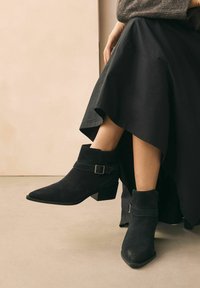 Botas de tobillo de ante negro con puntas afiladas, tacón de bloque y detalle de hebilla. Combinadas con una falda negra fluida. Textura lisa en todo.