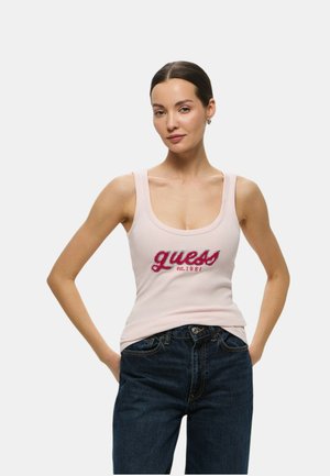 Top tank rosa chiaro con spalline larghe, con un logo "guess" rosso glitterato e il testo "est. 1981". Abbinato a jeans in denim scuro.