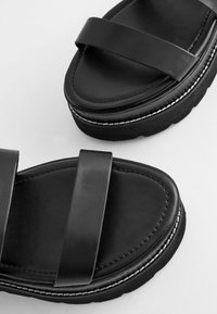 Melnas slidene sandals ar divām platām jostām, mīkstu zoli un teksturētu kāju pamatni ar kontrastējošu baltu šuvi pa malu.