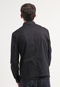 Musta takki, joka on valmistettu sileästä kankaasta, jossa on yksinkertainen kaulus, napitus ja suora leikkaus. Ei näkyviä koristeluja.