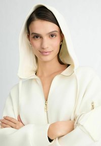 Zachte crèmekleurige hoodie met volledige rits, grote capuchon en een ritsvak op de mouw; de gladde textuur en losse pasvorm benadrukken het moderne design.