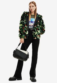 Giacca in pelliccia sintetica verde con dettagli rossi e crema, abbinata a una maglietta grafica, pantaloni neri e scarpe nere chunky. Include una borsa testurizzata.