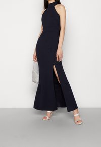 Vestido azul marino de cuello halter con una silueta ajustada y una abertura alta en el muslo. Combinado con un bolso plateado brillante y tacones con tiras. Textura suave.