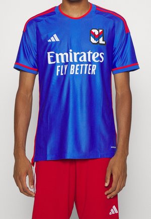 Maillot de sport bleu et rouge avec le logo "Emirates Fly Better" et l'emblème "OL" porté avec un short rouge, torse visible de la personne, mains détendues le long du corps.